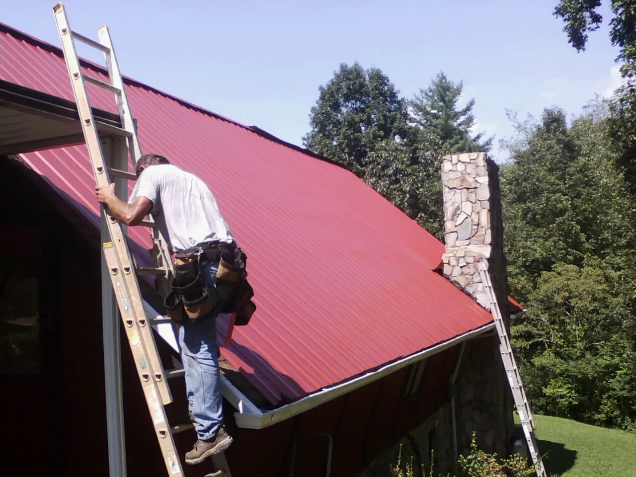 Roofer on a ladder beside a red metal roof and stone chimney — Soffit & Fascia Repair by Roofing Brothers in Hesperia